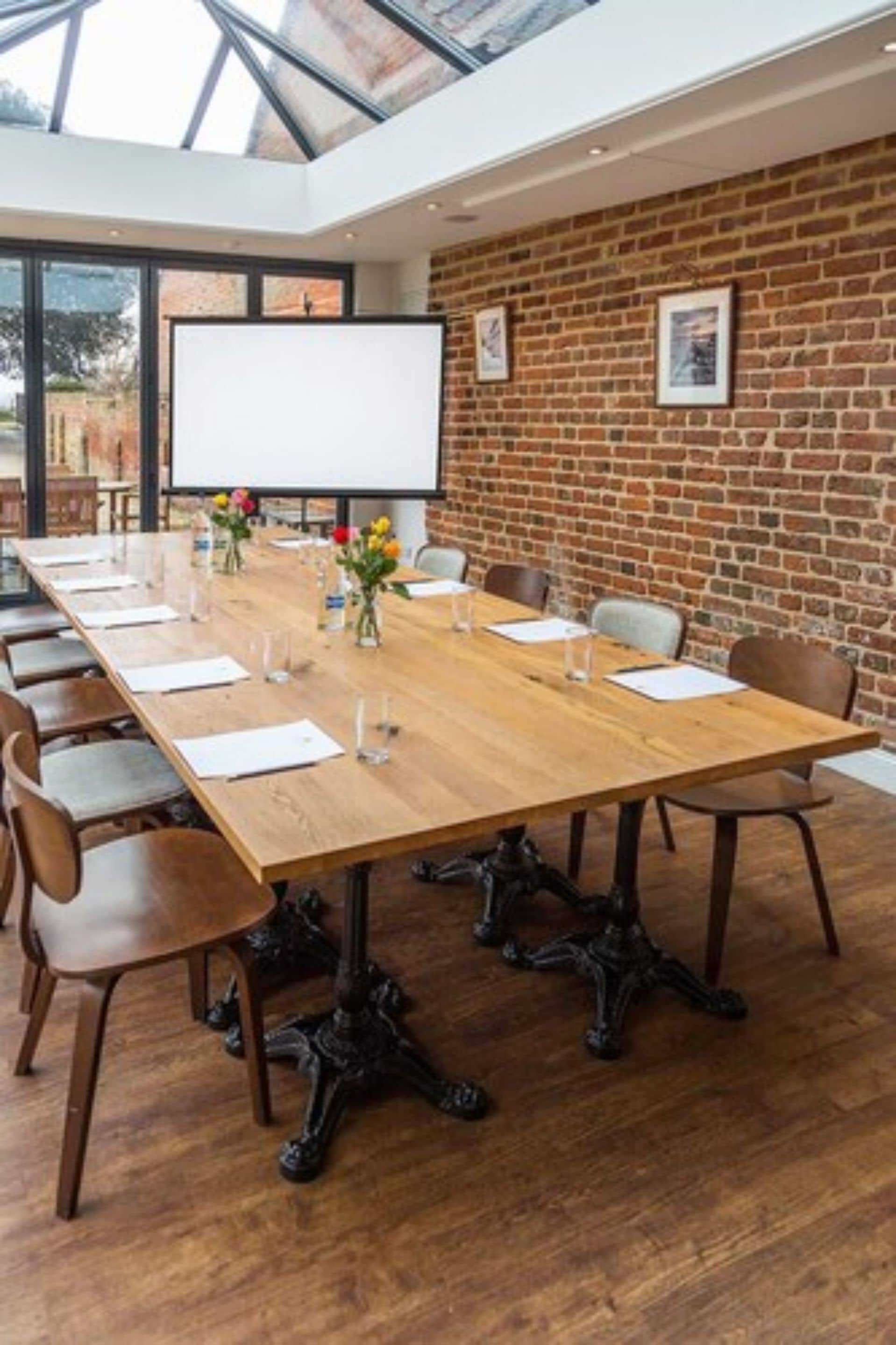 A conference room with a long wooden table, chairs, a projector screen, and a brick wall.