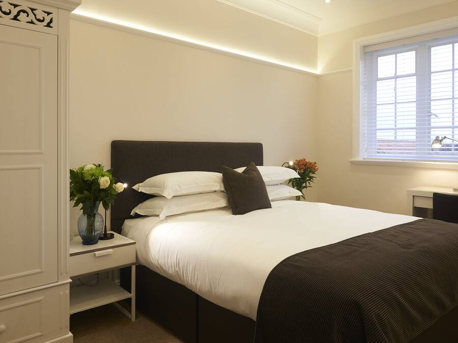 A neatly arranged bedroom with a large bed, bedside tables, a nightstand, a window with blinds, and a vase with flowers.