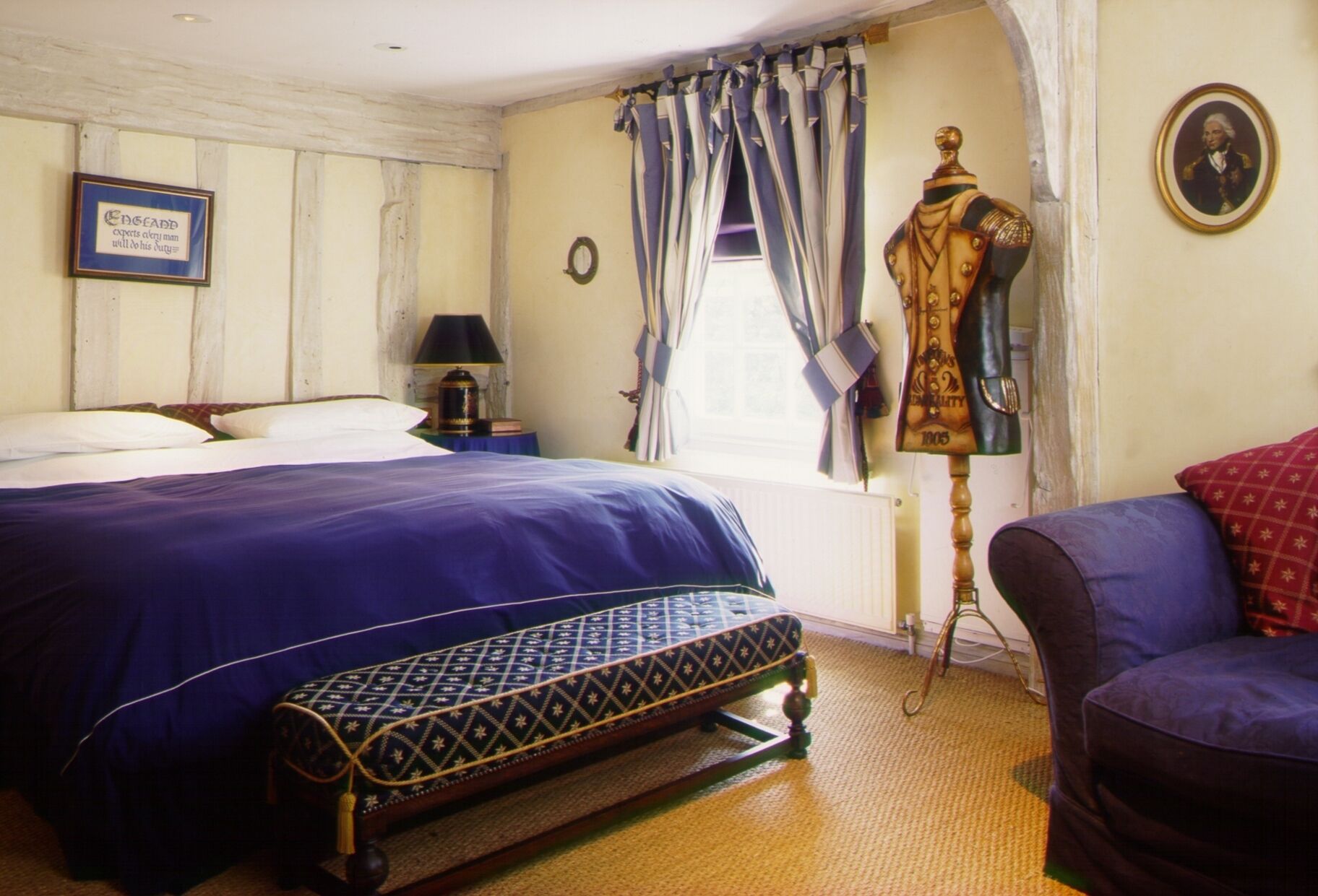 Een slaapkamer met een bed, een stoel, een lamp, een raam met gordijnen, een kapstok met een jas en een ingelijste citaat aan de muur.