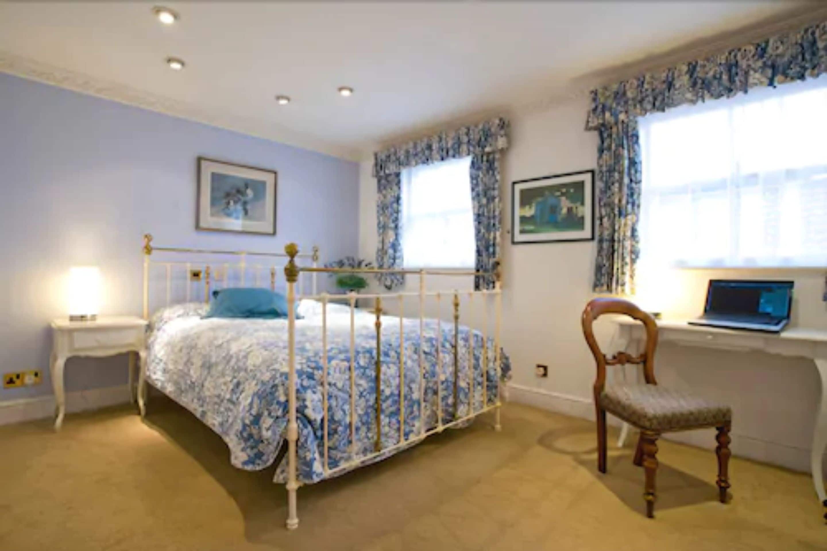 Une chambre à coucher avec un lit à baldaquin, une chaise en bois, un bureau avec un ordinateur portable et deux fenêtres ornées de rideaux à motifs floraux.