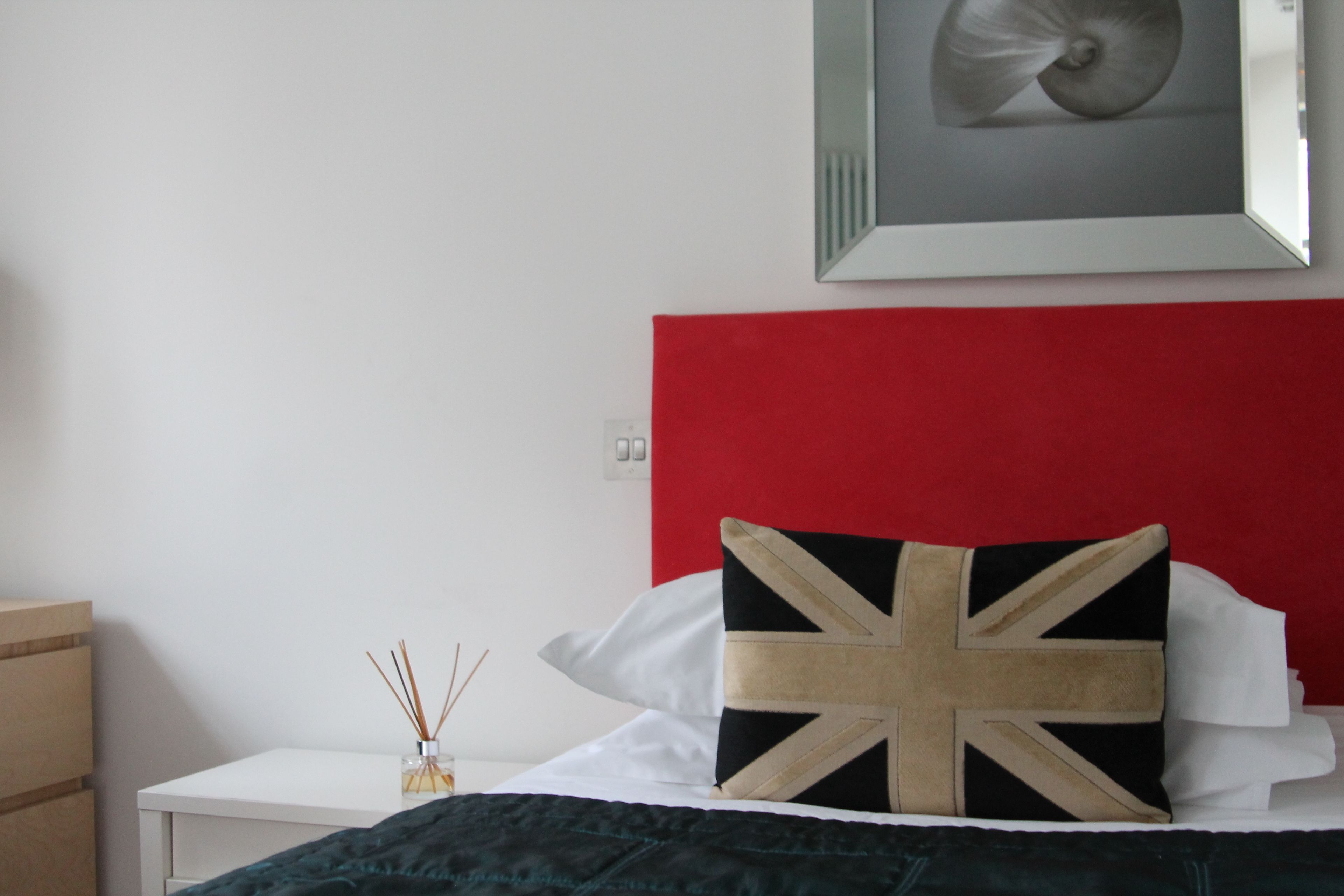 A neatly made bed with a Union Jack pillow, a white bedside table with a small vase, and a framed picture above the bed.
