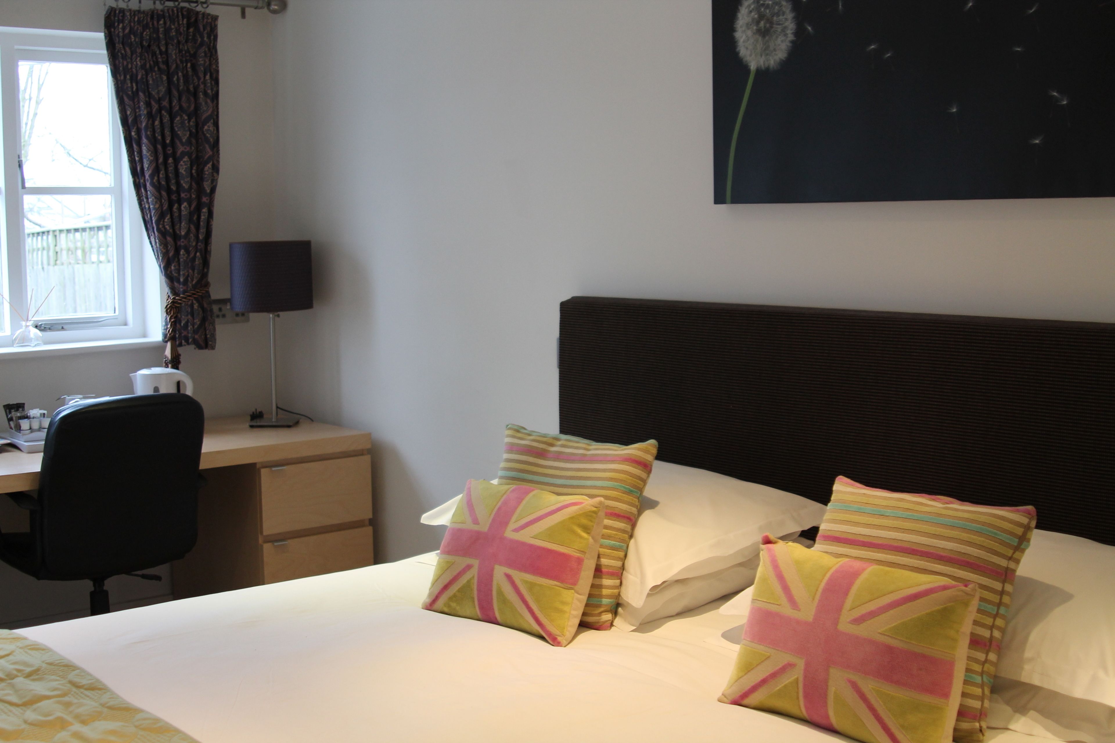 Una camera d'albergo con un letto, due cuscini con la Union Jack, una scrivania con una sedia, una lampada e una finestra con tende.