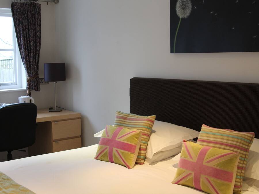 Una camera d'albergo con un letto, due cuscini con la Union Jack, una scrivania con una sedia, una lampada e una finestra con tende.