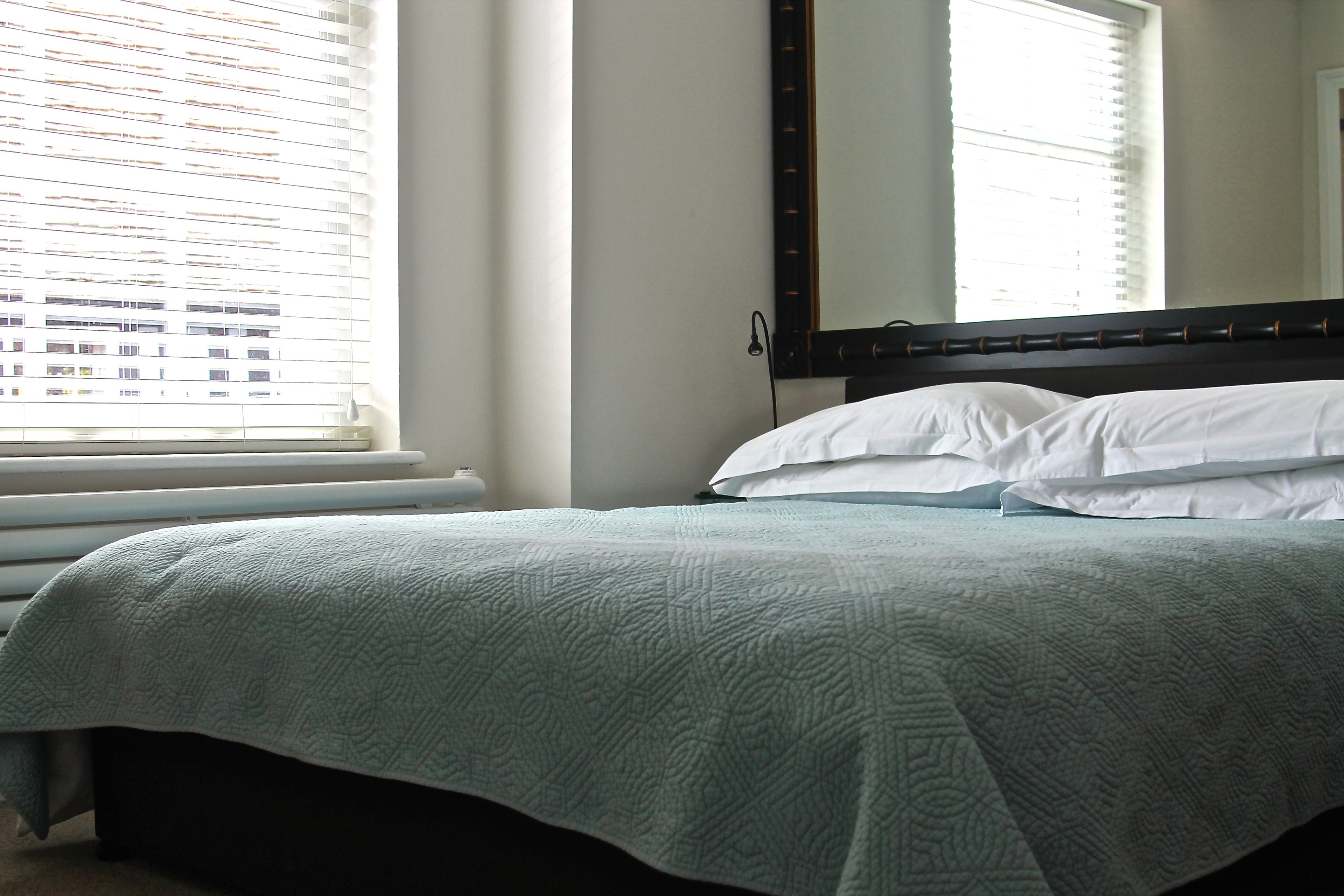 A neatly made bed with a quilt, a large mirror above it, and a window with blinds.