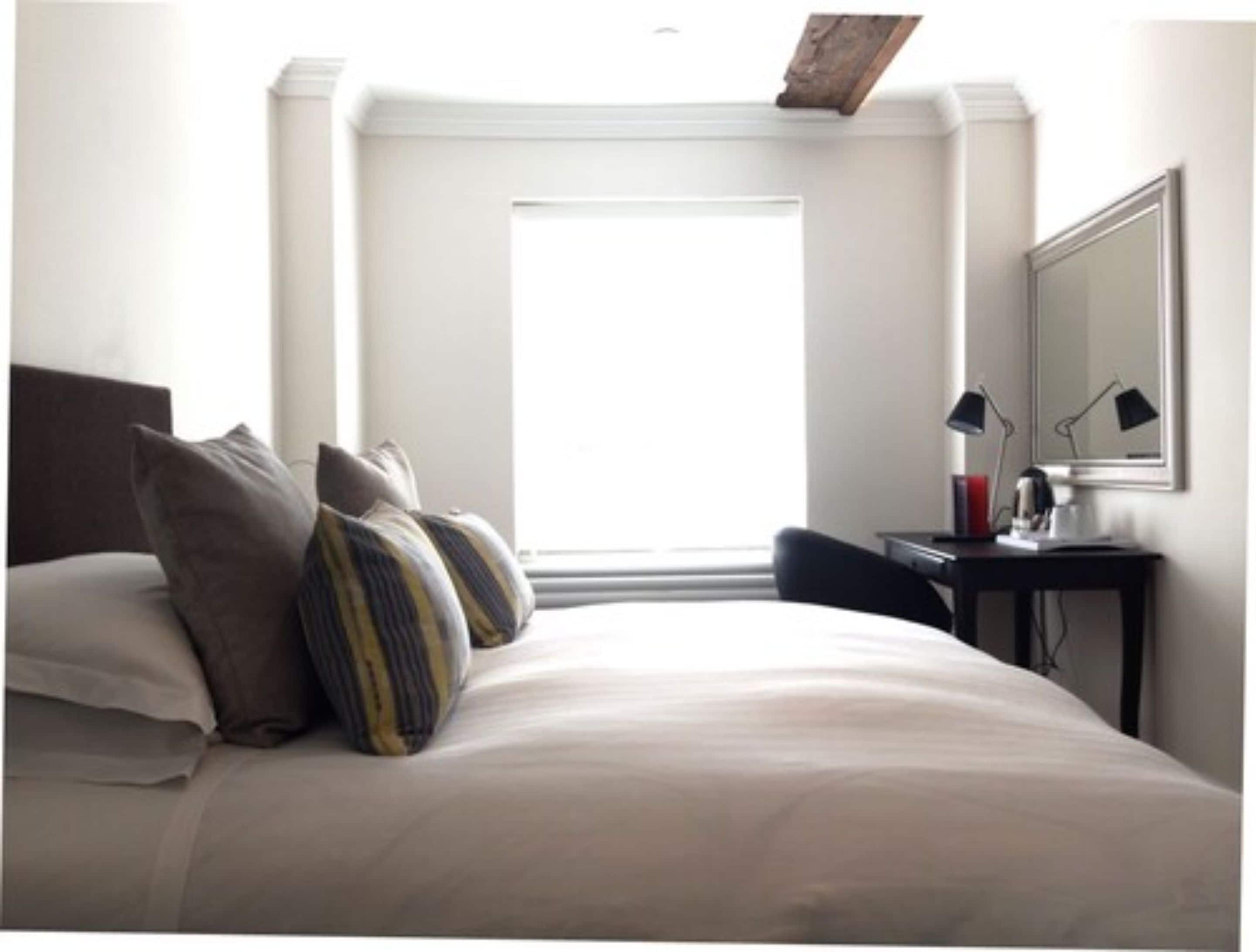 A hotel room with a bed, pillows, a desk with a lamp, and a window.
