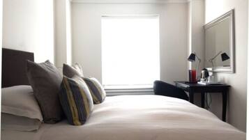 A hotel room with a bed, pillows, a desk with a lamp, and a window.