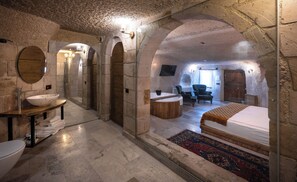 A stone-walled room with a large bed, a sitting area with chairs, a wooden table with a bowl sink, and a mirror. - Gedik Cave Hotel (Nevsehir)