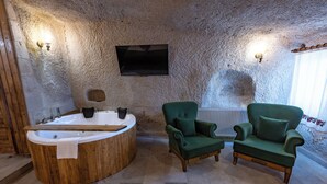Une pièce aux murs de pierre, avec une baignoire en bois, deux fauteuils verts et un téléviseur à écran plat fixé au mur.