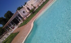 Blick auf einen Poolrand an einem modernen Gebäude, mit Pool in gebogener Form, Steinmauern und einem gepflegten Rasen.