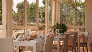 A sunlit dining area with a table set for breakfast, surrounded by wicker chairs, and large windows offering a view of trees and a garden. - B&B La Vigna (Carovigno)