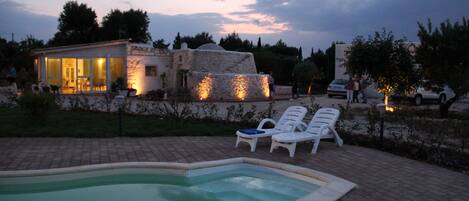 A swimming pool with lounge chairs, a stone building with a glass facade, and a sunset sky.