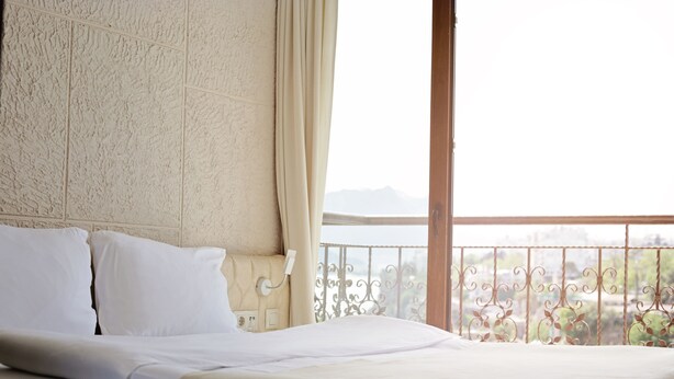 A bed with white linens in a room with a textured beige wall and a view of a balcony with a metal railing. - Patron Hotel (Antalya)