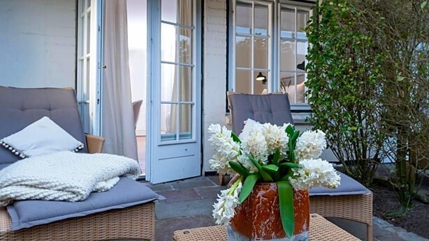 Ein gemütlicher Sitzbereich im Freien mit Korbmöbeln, einer weißen Vase mit Blumen und einer Tür zu einem angrenzenden Raum.