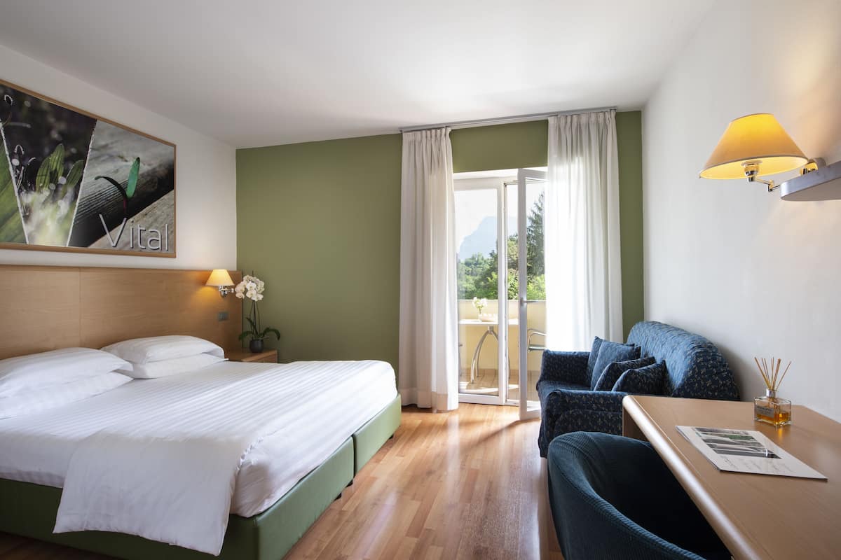 A hotel room with a bed, a desk, a chair, a lamp, and a view of the outdoors.