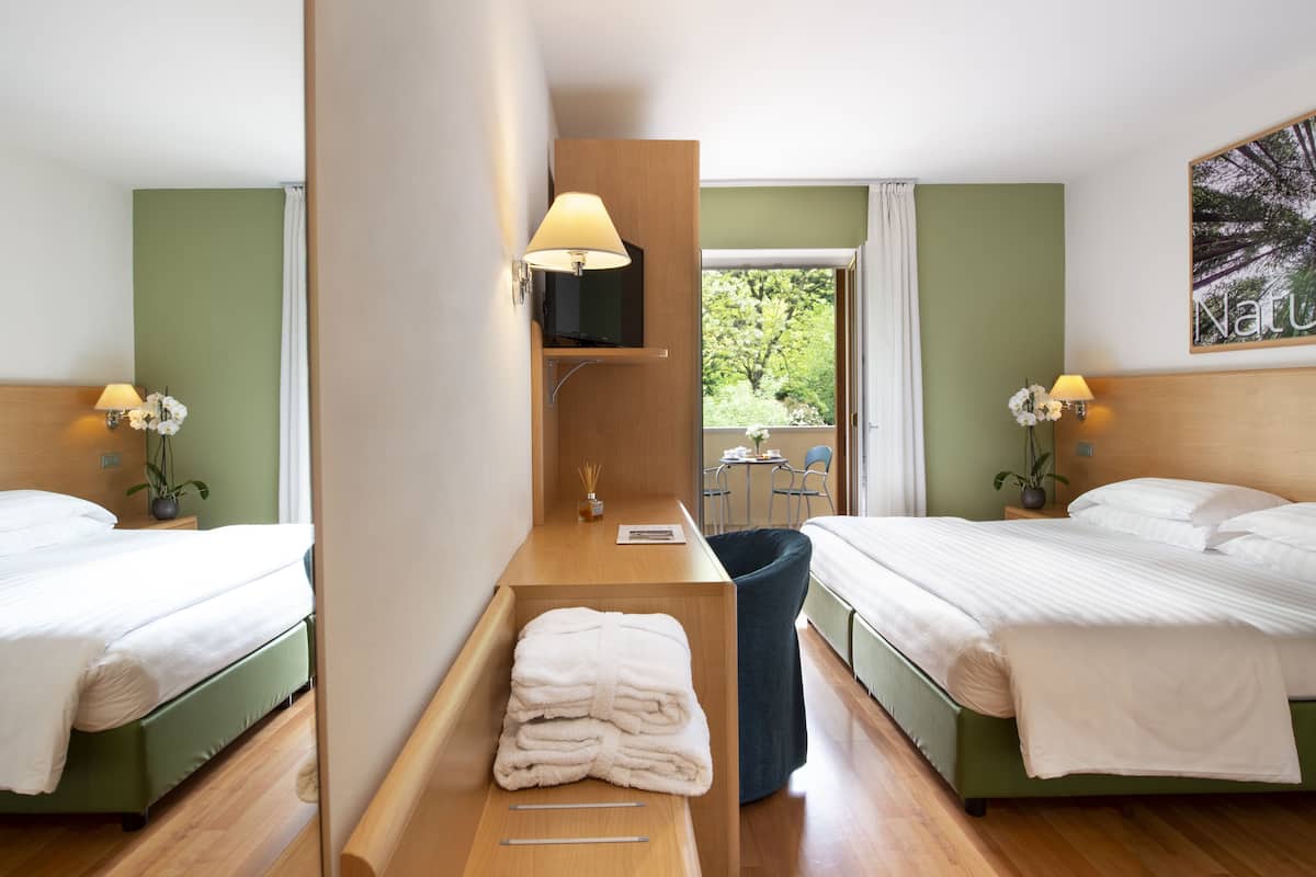 A hotel room with two beds, a wooden desk, a small table, and a view of greenery outside.