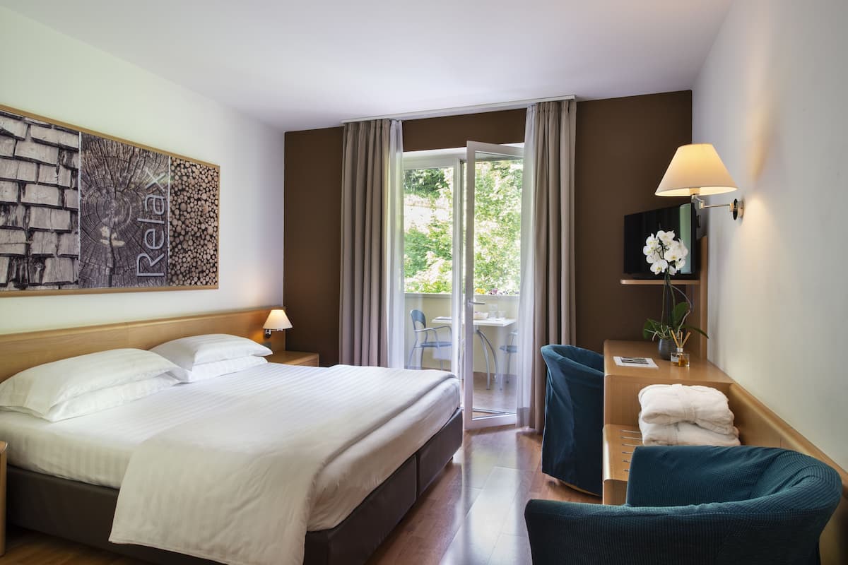 A hotel room with a bed, a desk with a lamp, a chair, a small table with a plant, and a sliding glass door leading to an outdoor area.
