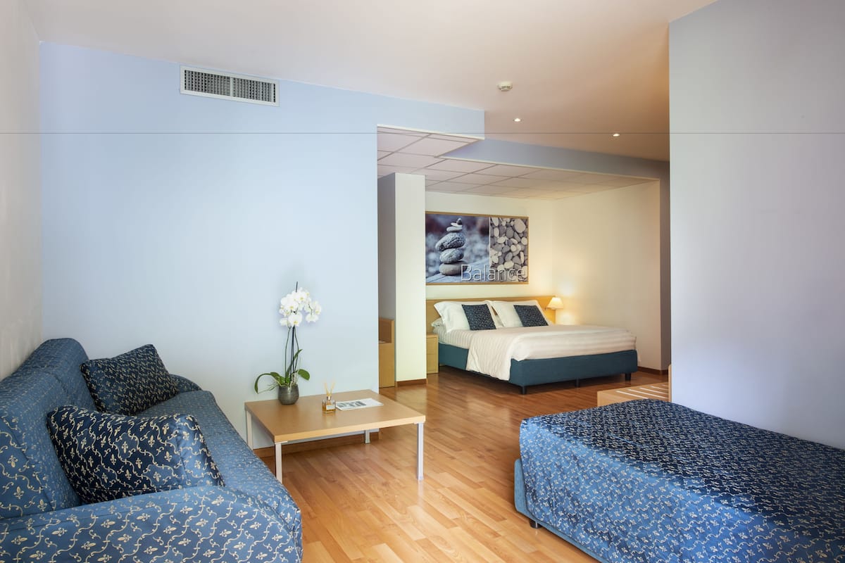 A modern hotel room with a blue sofa, a small table with a vase, and a bed with a headboard.
