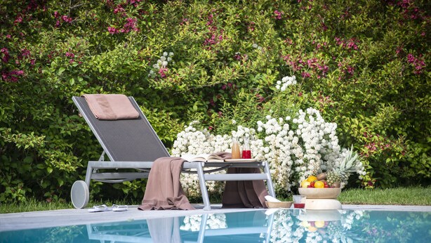 Una silla de descanso junto a la piscina, con una toalla, un libro y un frutero.