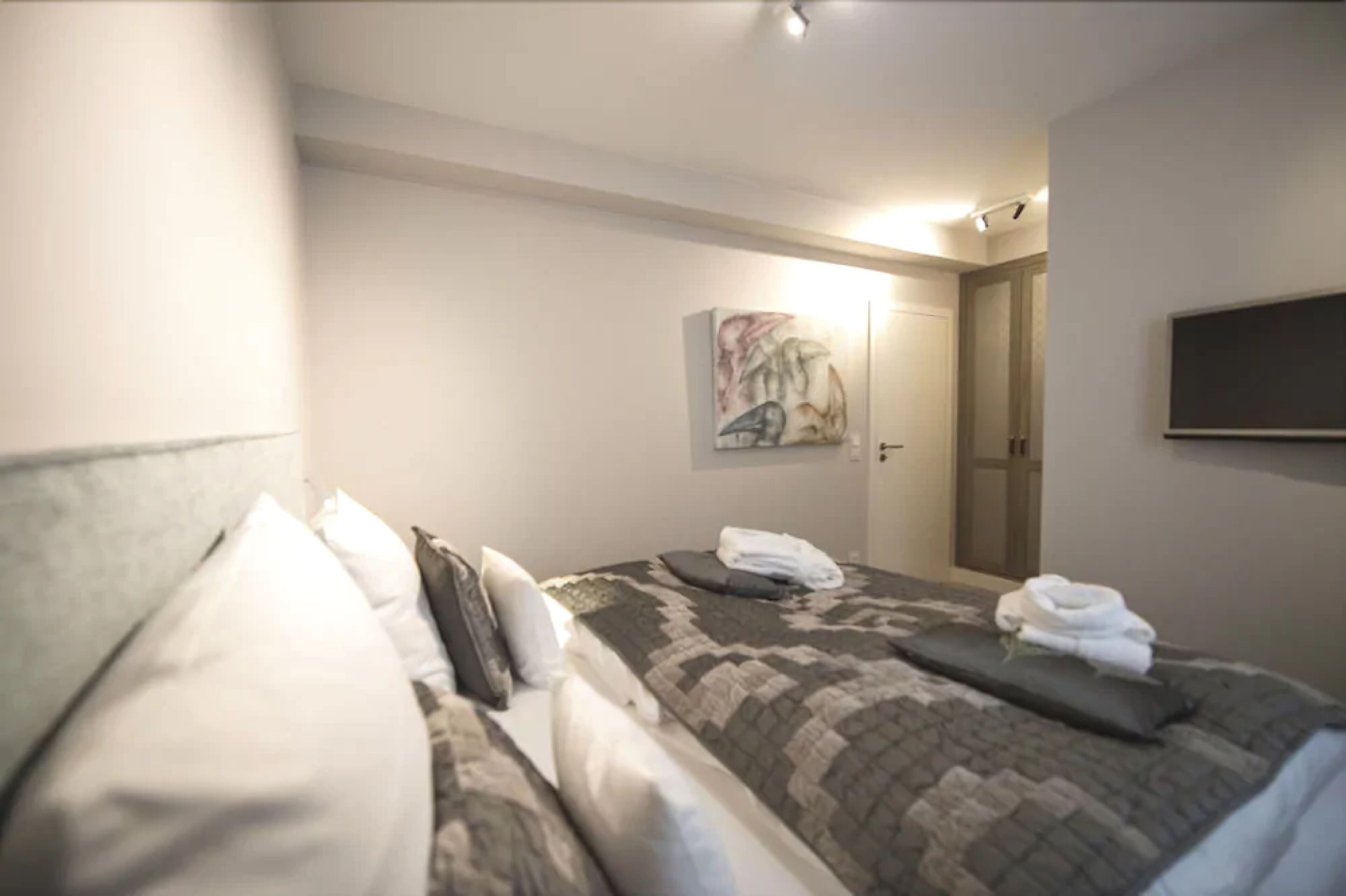 A hotel room with a bed, pillows, a folded towel, a painting on the wall, and a door.