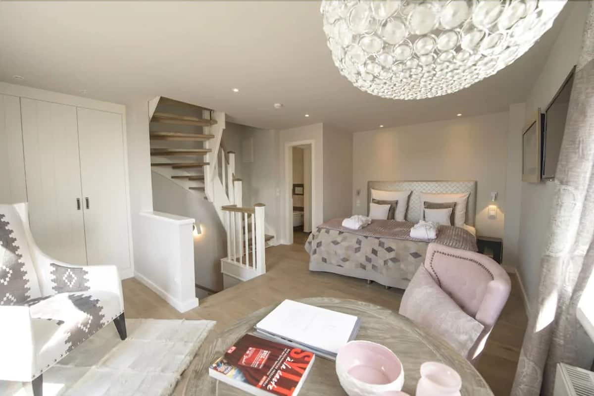 A bedroom with a bed, a chair, a table with books, a staircase, and a chandelier.
