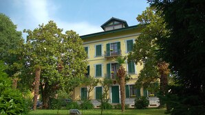 A two-story building with green shutters, a balcony, and a garden with trees and plants. - Hotel Damian (Trofarello)