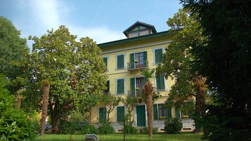 Um edifício de dois andares com persianas verdes, varanda e jardim com árvores e plantas.