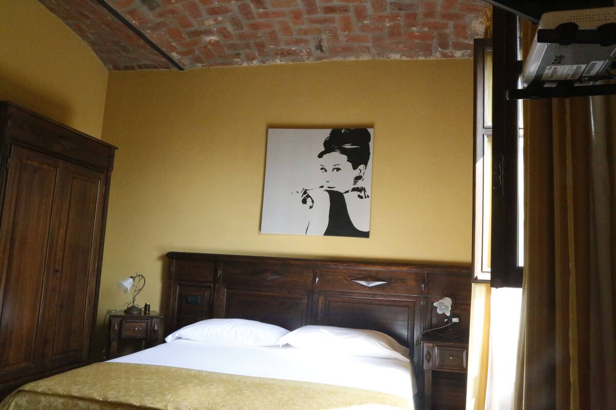Una habitación de hotel con cama, cabecero de madera, mesita de noche, lámpara y un cuadro en la pared.