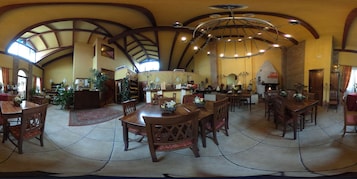Uma sala de jantar ampla, com pé direito alto e vigas de madeira, com várias mesas e cadeiras.