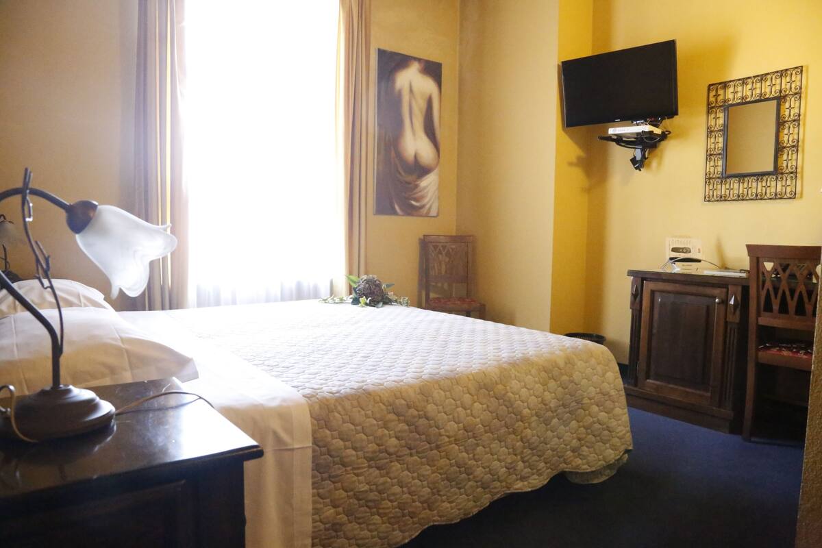 Habitación de hotel con una cama, un escritorio con lámpara, un televisor montado en la pared y un espejo.