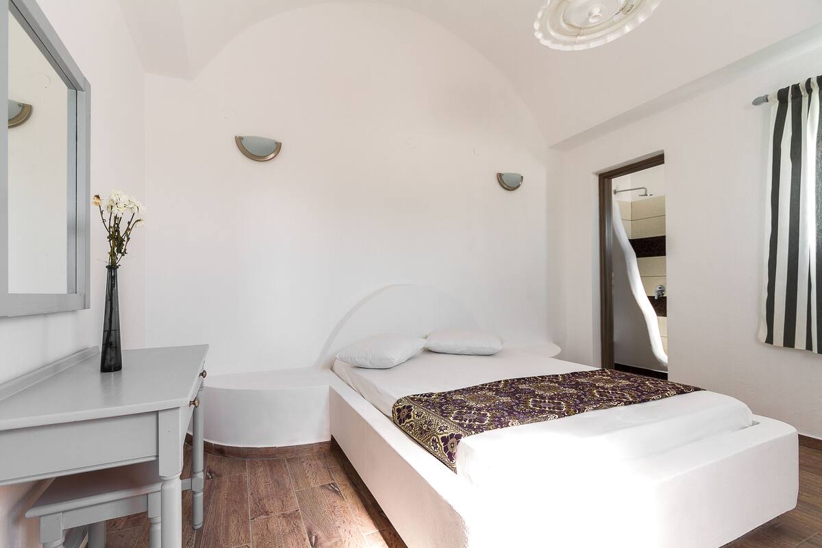Ein modernes Schlafzimmer mit einem weißen Bett, einem weißen Schreibtisch und einem durch einen Bogen sichtbaren Badezimmer.