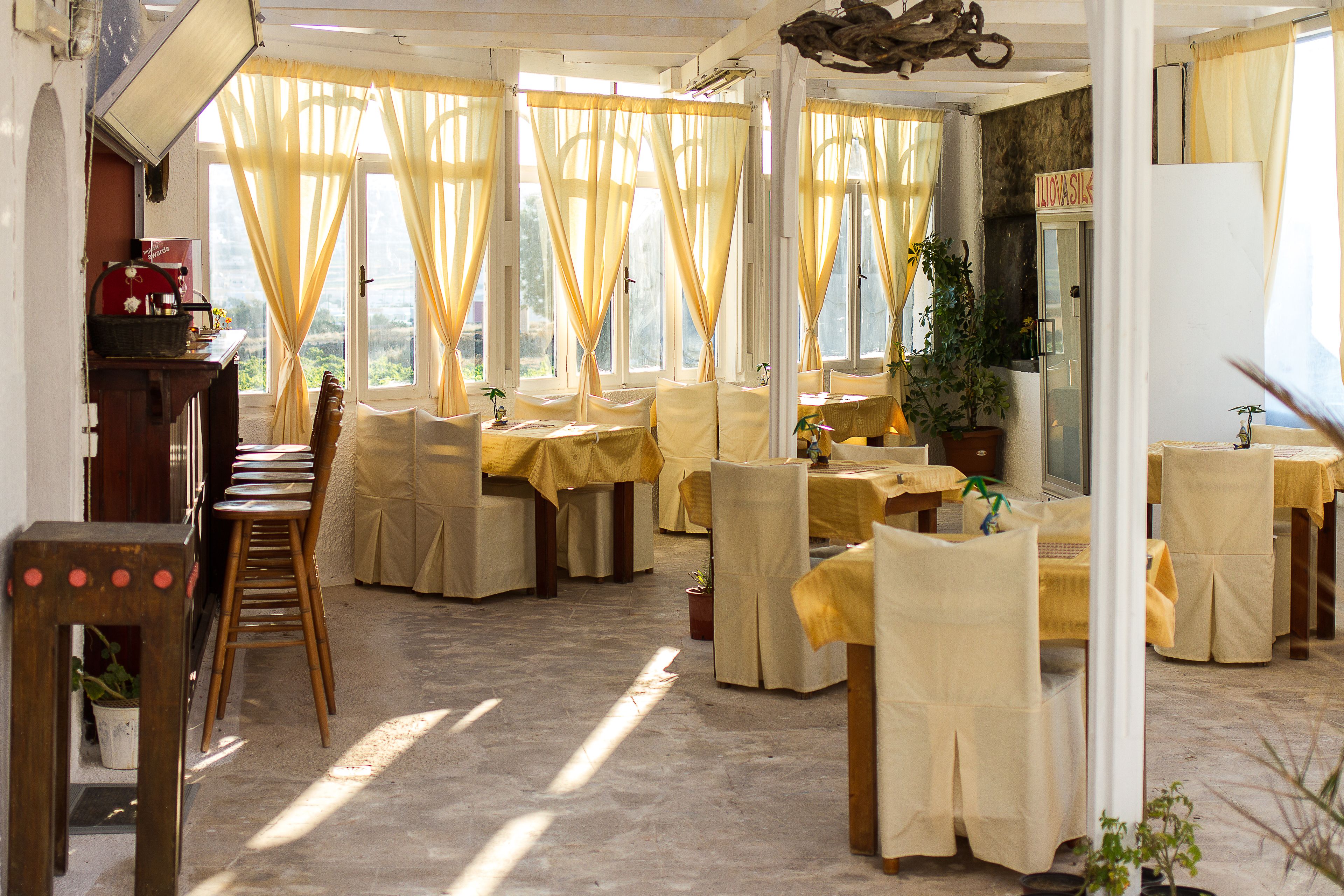 Un espace repas avec des tables et des chaises recouvertes de nappes jaunes, de grandes fenêtres avec des rideaux et une vue sur l’extérieur.