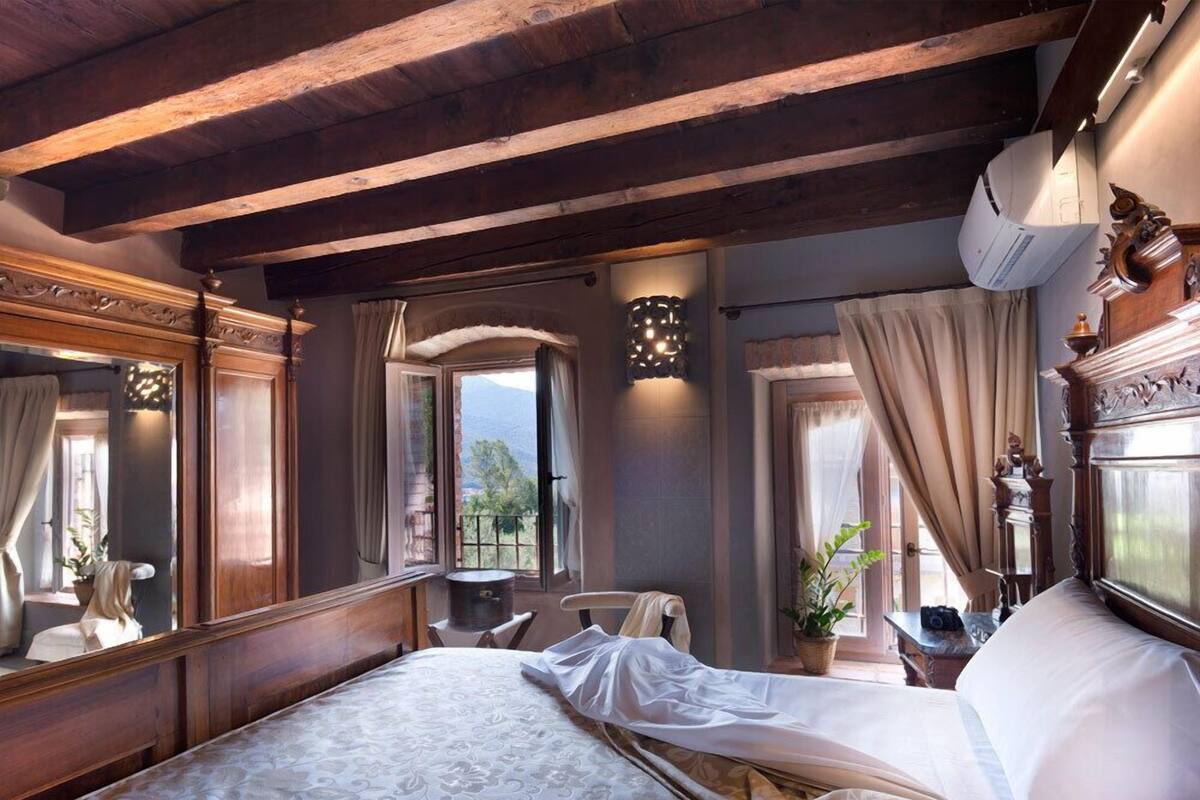 A bedroom with a large bed, wooden beams, a window with a view, a bedside table, and a mirror.