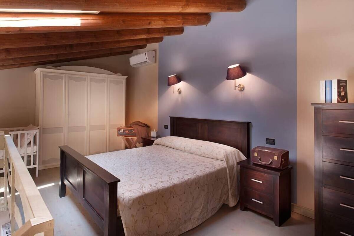 A bedroom with a wooden ceiling, a bed with a patterned bedspread, two wall-mounted lamps, a wardrobe, and a chest of drawers.