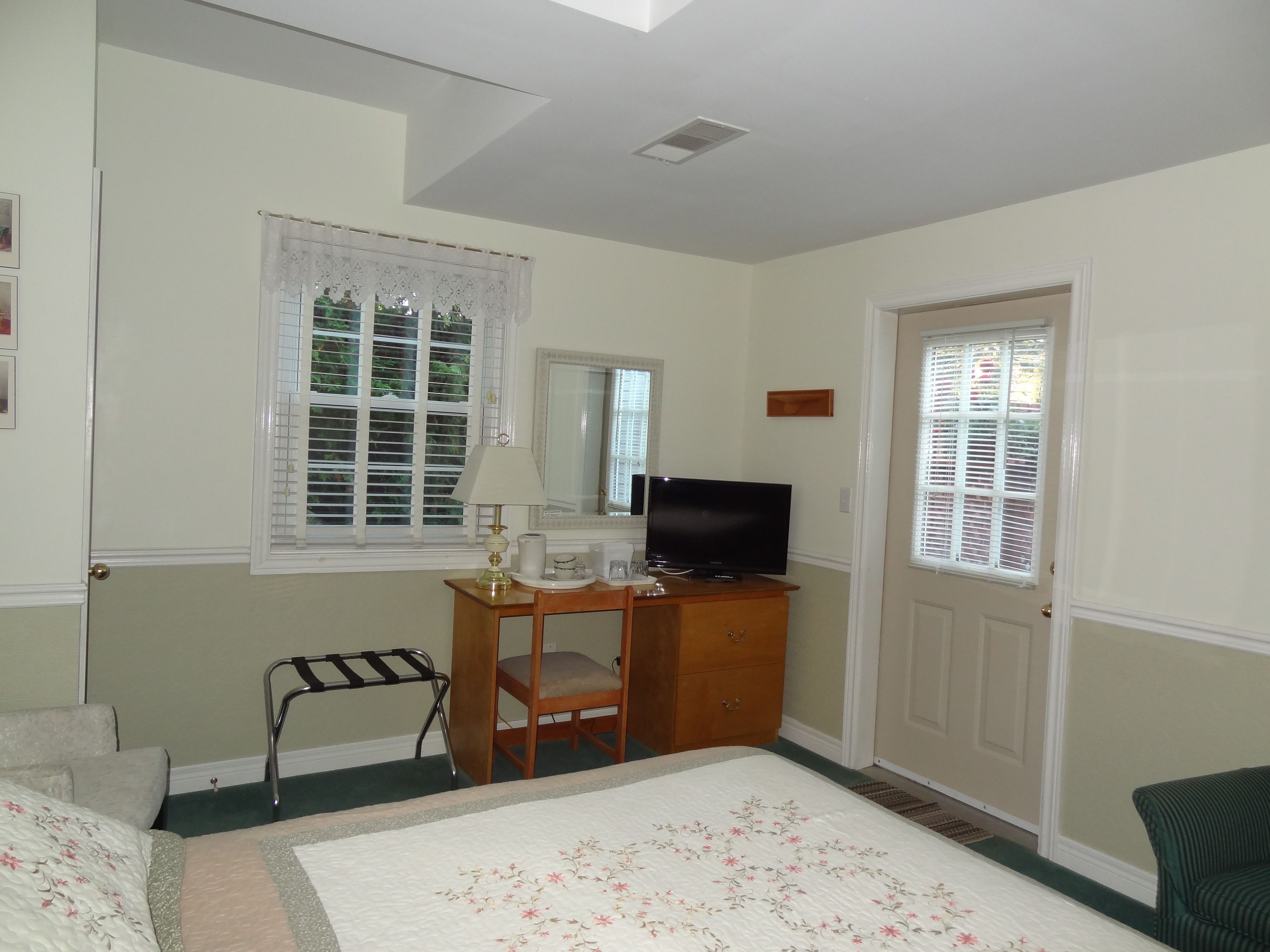 A bedroom with a bed, a desk with a television, a chair, a window with blinds, and a door.
