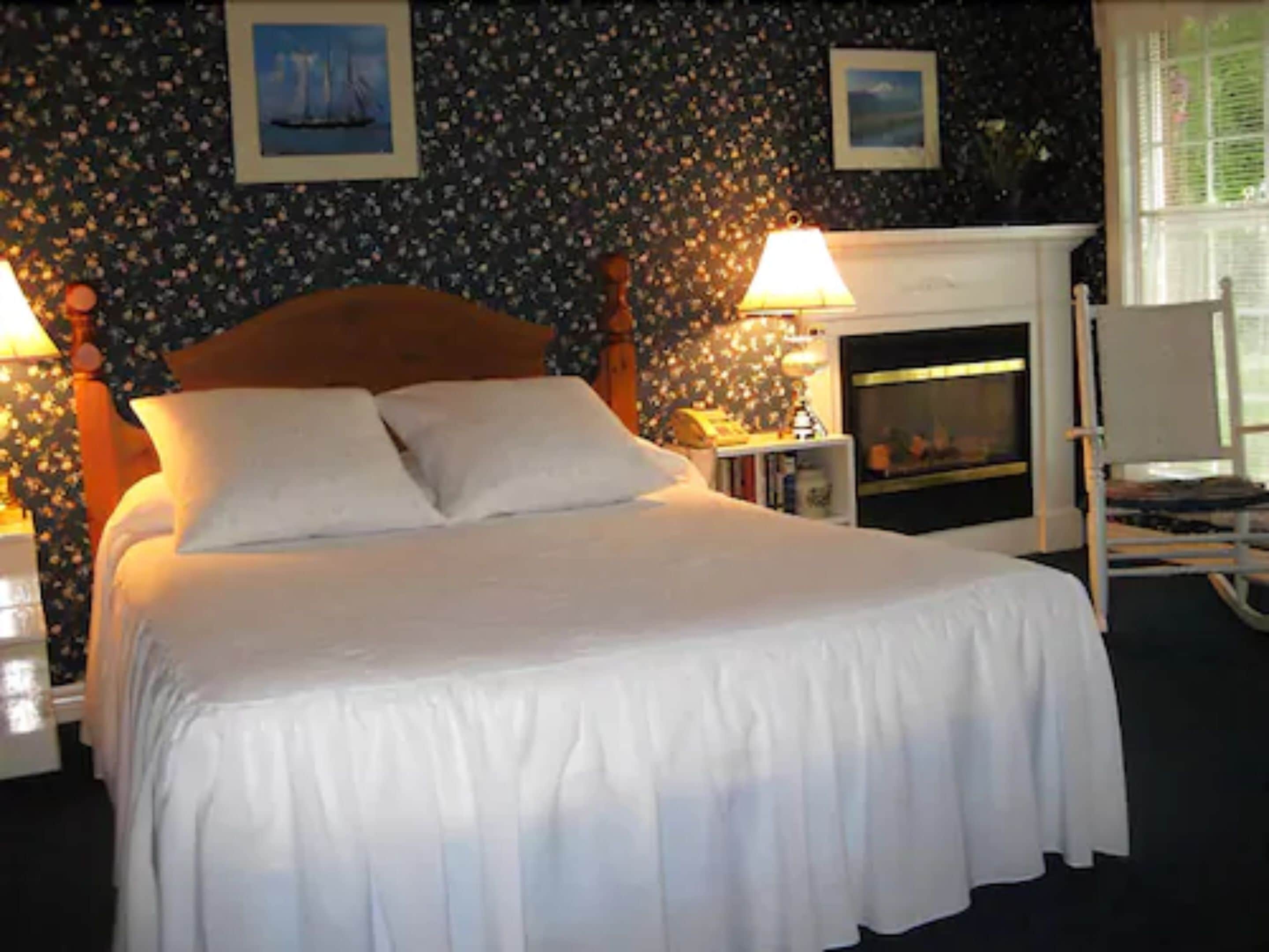 Una cama bien hecha con sábanas blancas en una habitación con papel tapiz floral, dos lámparas de noche y una chimenea.