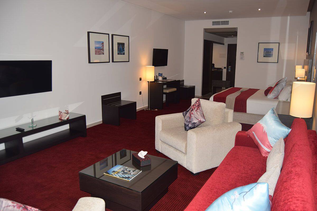 A modern hotel room with a red carpet, a sofa, a chair, a coffee table, a TV, and a desk with a lamp.