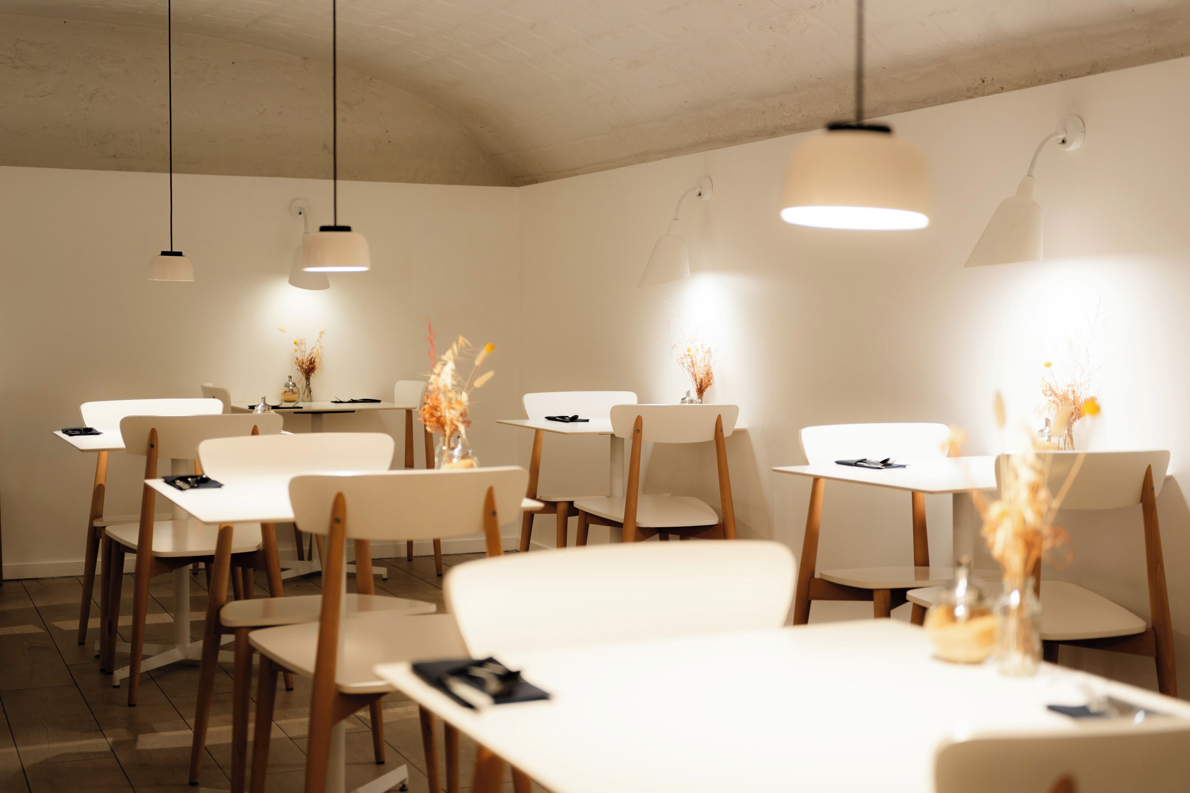 A dining area with white tables and chairs, overhead lighting, and decorative vases with dried flowers.