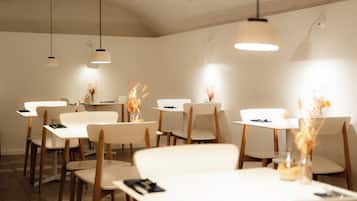 A dining area with white tables and chairs, overhead lighting, and decorative vases with dried flowers.