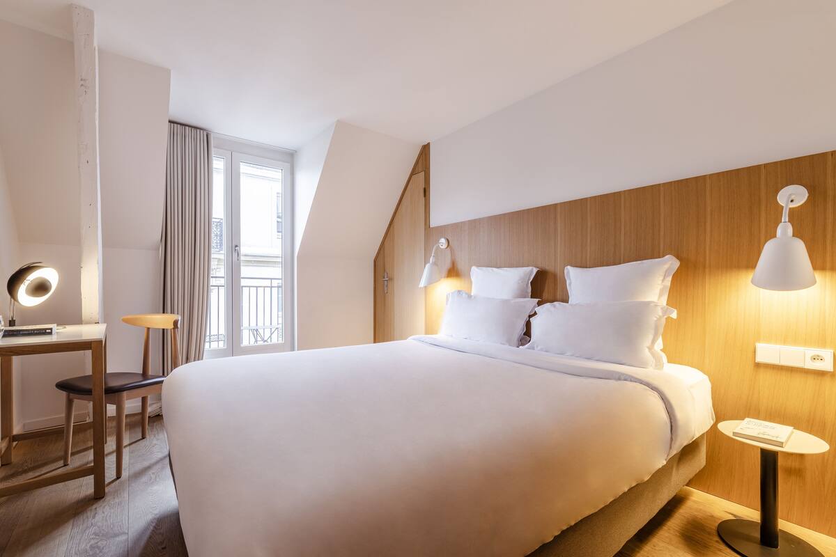 A modern hotel room with a large bed, a desk, and a chair. There is a window with curtains, a wooden headboard, and a wall-mounted lamp.