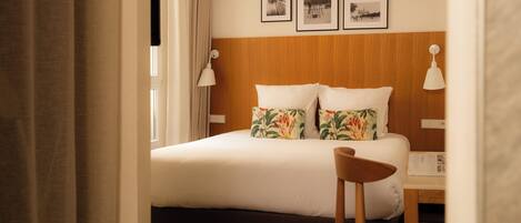 A hotel room with a bed, two pillows, a nightstand, a chair, and a window with curtains. There are three framed pictures on the wall above the bed.
