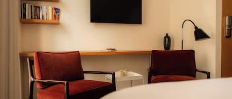 A hotel room with two red upholstered chairs, a small white table, a black floor lamp, a flat-screen TV on the wall, and a bookshelf with books.