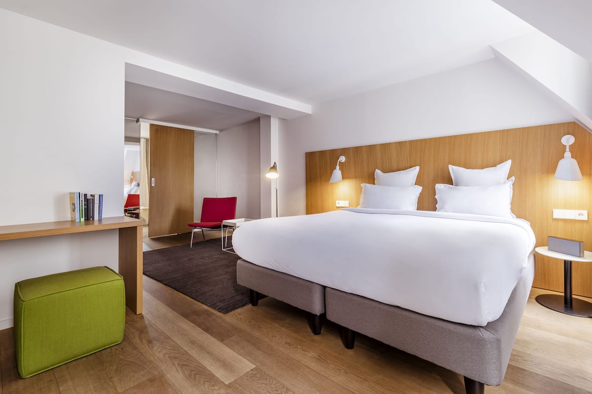 A modern hotel room with a large bed, a wooden headboard, a green ottoman, a desk with a chair, and a small side table with a lamp.