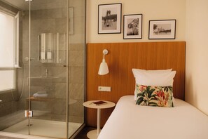 A hotel room with a glass shower enclosure, a wooden headboard, a bedside table with a book, and three framed photographs on the wall.