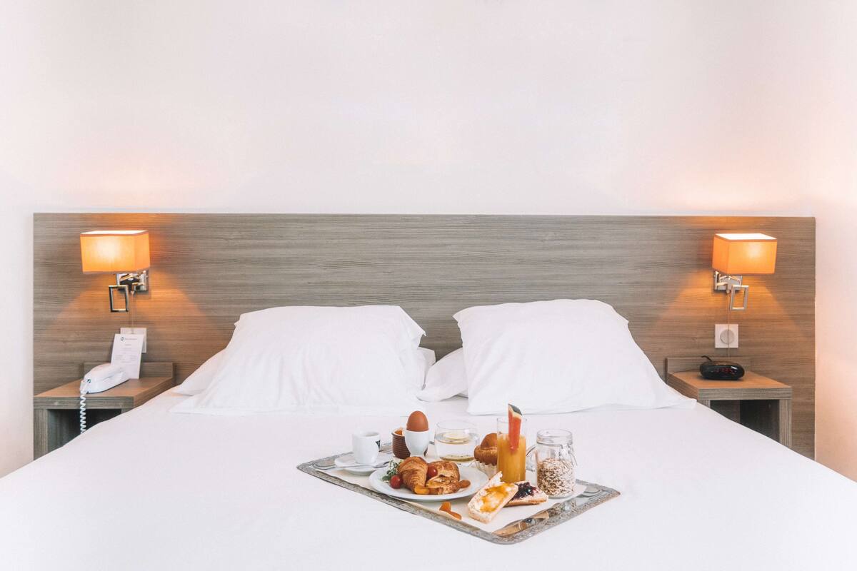 Une chambre d’hôtel comprenant un lit, deux tables de chevet, un plateau avec des articles pour le petit-déjeuner et deux lampes fixées au mur.