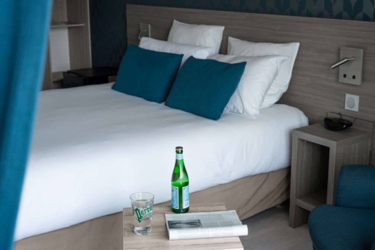 Un lit bien fait, avec des oreillers blancs et turquoise, une bouteille d’eau Perrier et un verre posés sur une table de chevet en bois.