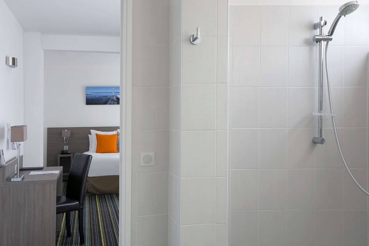 Une chambre d’hôtel avec une douche, un lit avec un oreiller orange, un bureau avec une lampe et un tableau au mur.
