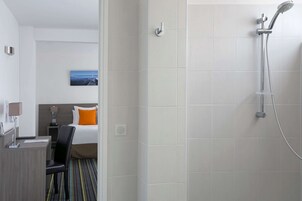 A hotel room with a shower, a bed with an orange pillow, a desk with a lamp, and a picture on the wall.