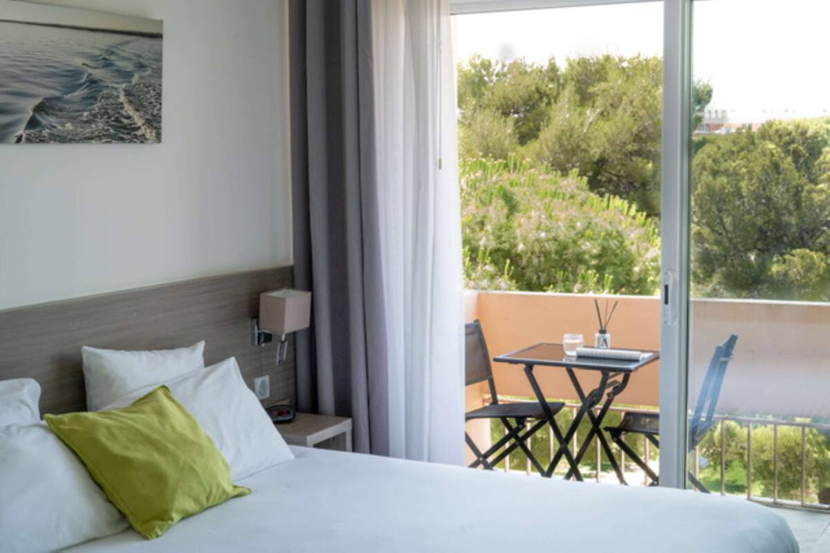 Une chambre d’hôtel avec un lit, une table de chevet, une lampe et une vue sur un balcon équipé d’une table et de chaises.