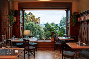 A restaurant with wooden tables and chairs, a large window with a view of greenery and mountains, and a bookshelf with books and plants. - La Maison d'Igor (Morges)