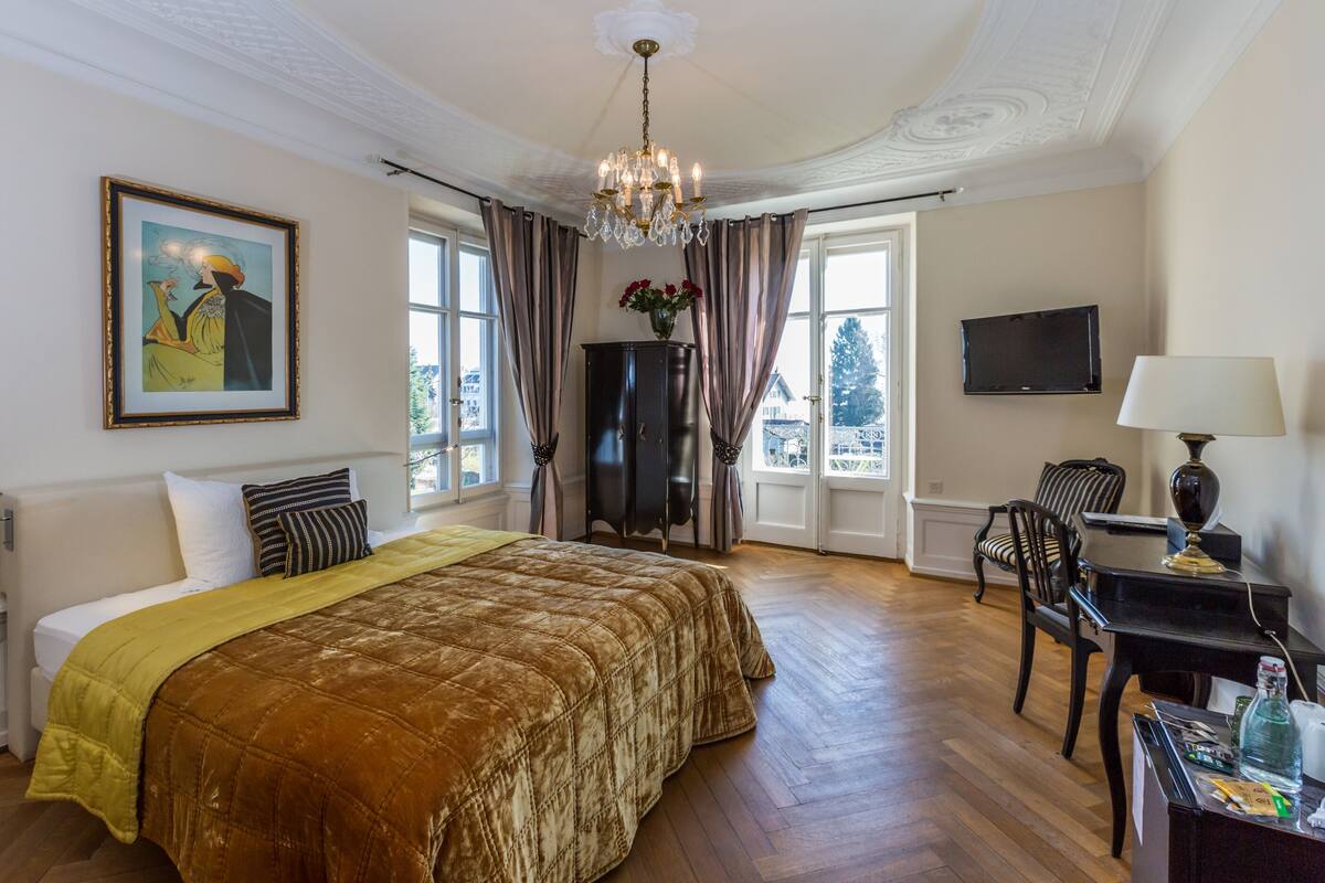 A hotel room with a large bed, a chandelier, a TV, a desk with a lamp, and a painting on the wall.